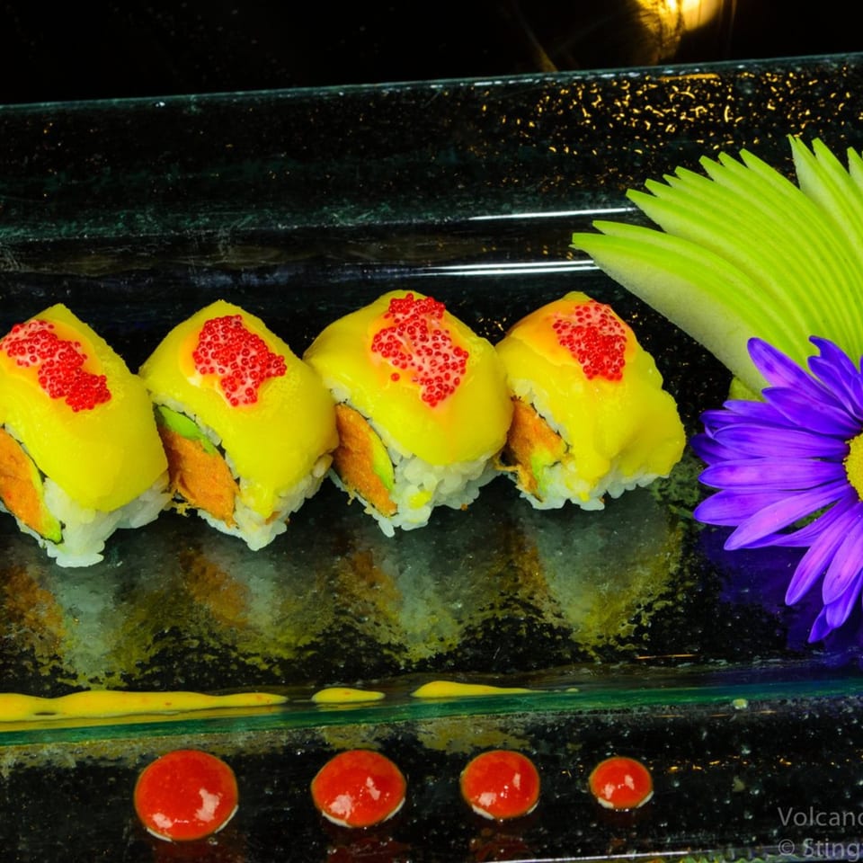 Four sushi rolls with yellow topping and red caviar, garnished with a purple flower and green leaves.