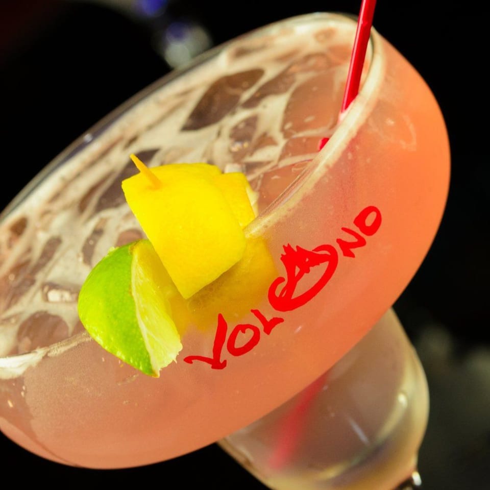 Pink cocktail with ice, a red straw, and citrus garnishes in a glass with "VOLCANO" in red.
