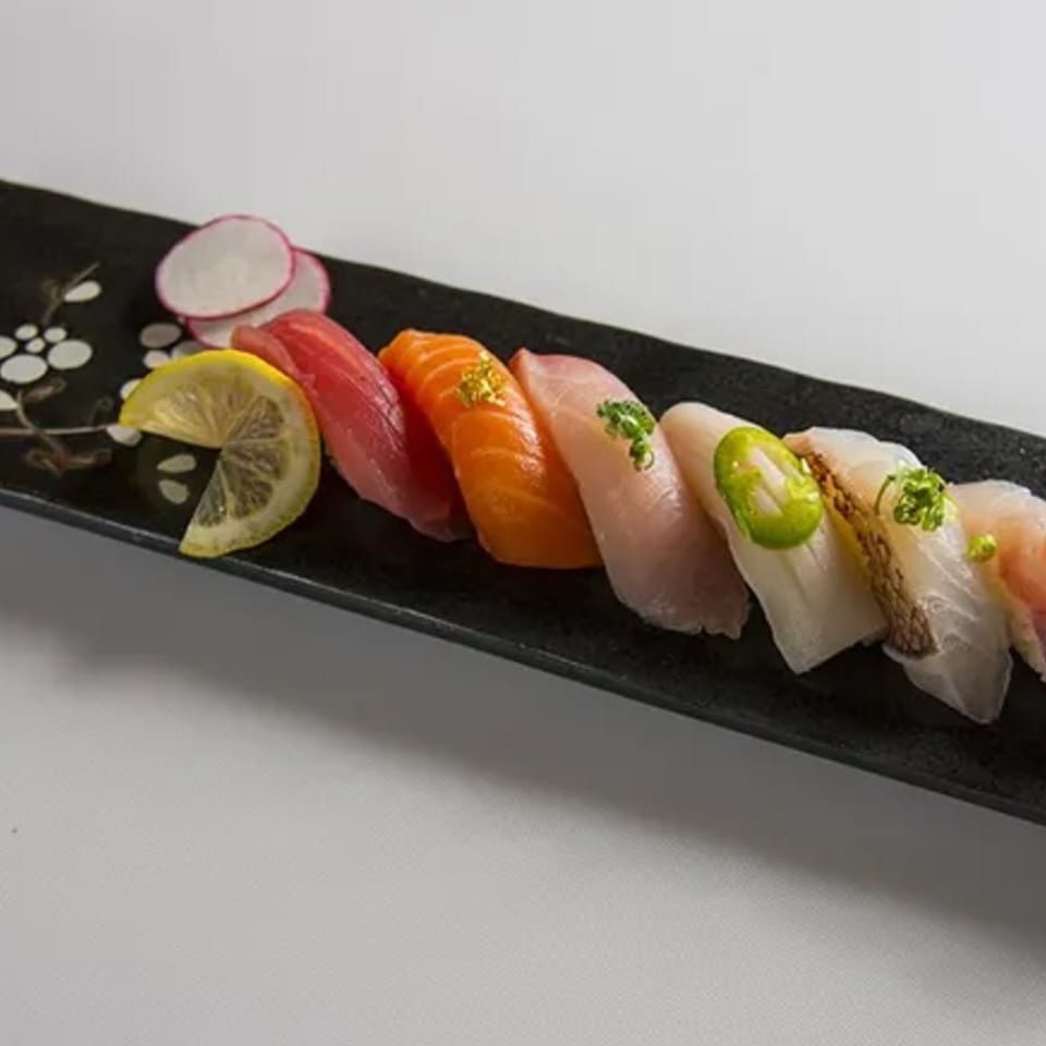 A black rectangular plate holds various colorful nigiri sushi pieces, garnished with lemon and radish.